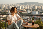 a brazilian woman, with short bob, she is wearing a white t-shirt and jeans, she is seated on a balcony we see a city behind her. On the table there is 2 glasses and a bottle of wine